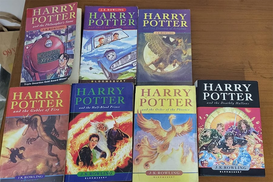 Seven Harry Potter books with illustrated covers arranged on a table, showing titles from “The Philosopher’s Stone” to “The Deathly Hallows.” Each cover features different artwork related to the story.