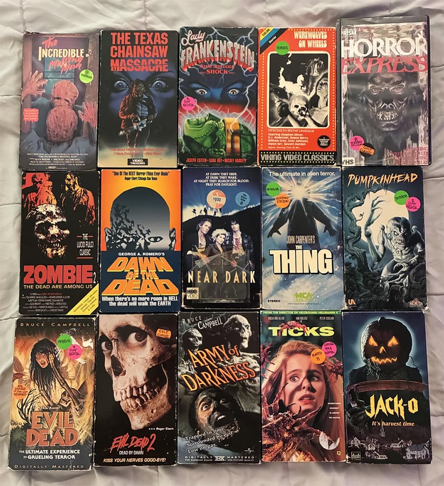 Fifteen vintage horror VHS tapes are arranged in three rows. Titles include “The Evil Dead,” “The Thing,” “Pumpkinhead,” “Army of Darkness,” and more, each with colorful cover art and price stickers.