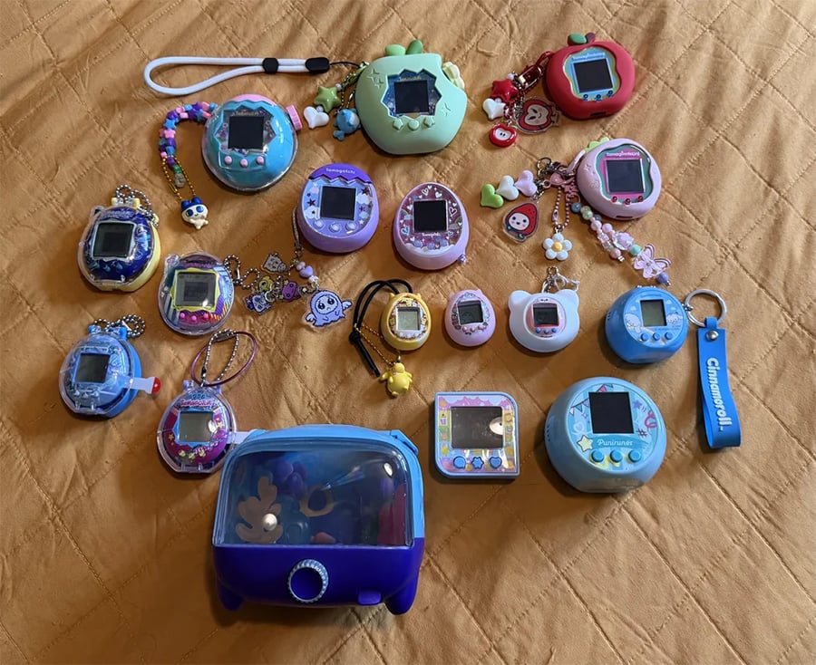 A collection of colorful Tamagotchi devices with keychains and charms arranged on a tan quilted surface, including unique shapes like a TV and designs with cartoon characters.
