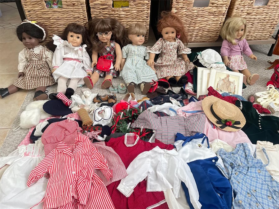 Six dolls are seated in a row on the floor, surrounded by various doll clothes, hats, and accessories. Behind them are woven baskets. The scene looks like a doll collection display.