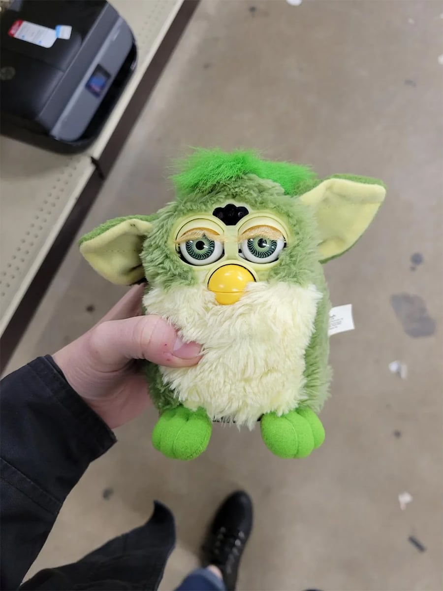 A person holds a green and yellow Furby toy with droopy eyes in a store, standing on a beige floor with shelves in the background.