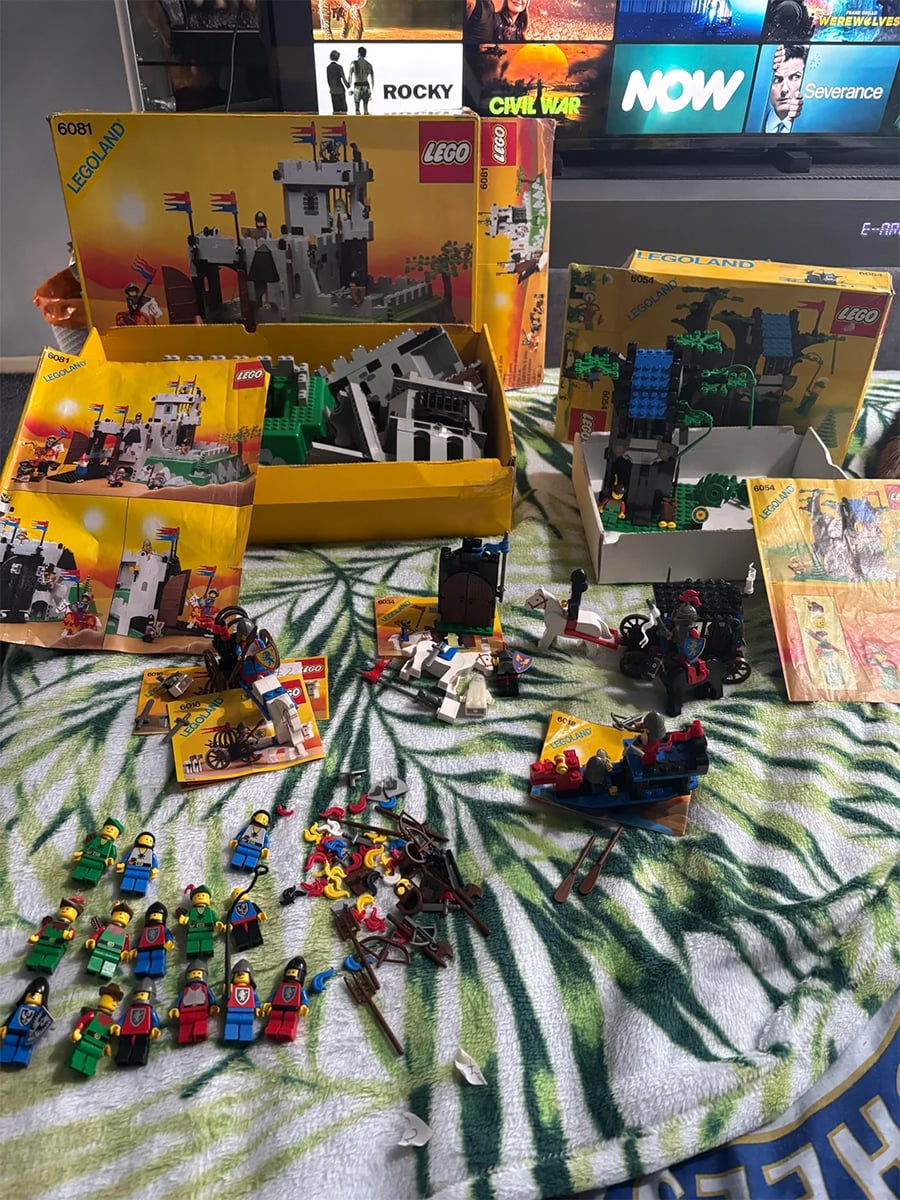 LEGO castle sets, minifigures, and instructions are spread out on a leaf-patterned cloth. LEGO boxes and assembled structures are visible, with TV shows displayed on a screen in the background.