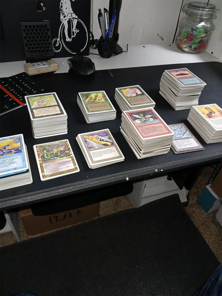 Stacks of Magic: The Gathering cards are organized on a black desk, with a keyboard, mouse, and glass jar in the background. The desk is in a room with shelving and a bike illustration on the wall.