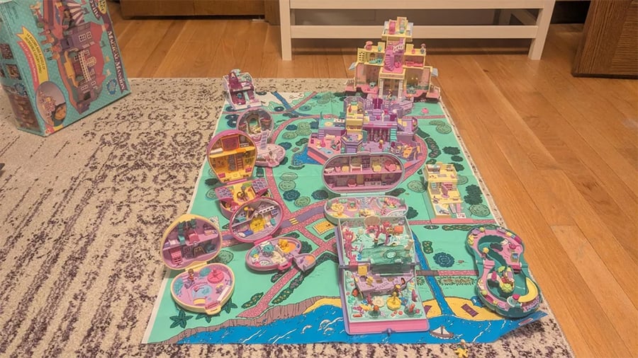 Toy playsets arranged on a colorful mat with roads and buildings, featuring pastel-colored miniature houses and figures. The scene is set indoors on a wooden floor next to a patterned rug.