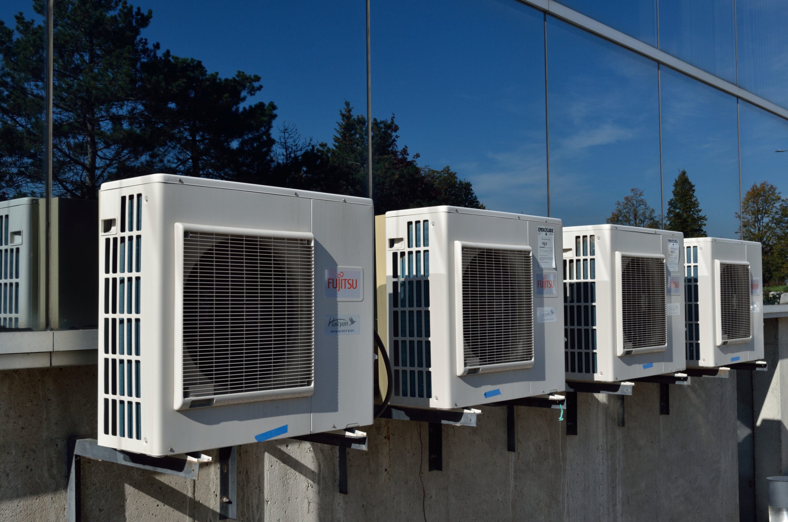 Four outdoor air conditioning units mounted on a concrete wall, reflected in nearby glass windows, with trees and a blue sky in the background.