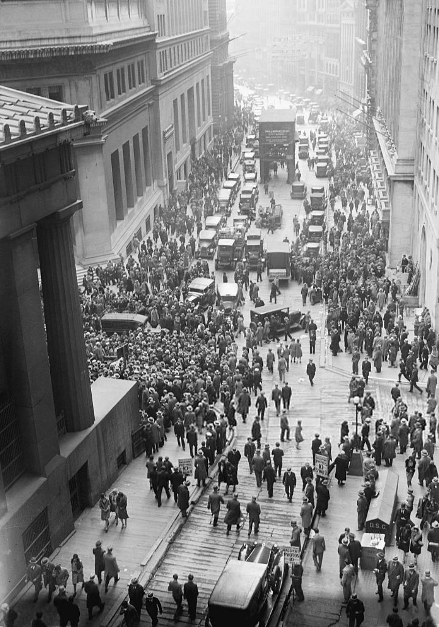 Black and white photo of a crowded city street filled with people and cars, likely from the early 20th century, with tall buildings lining both sides and a bustling, busy atmosphere.
