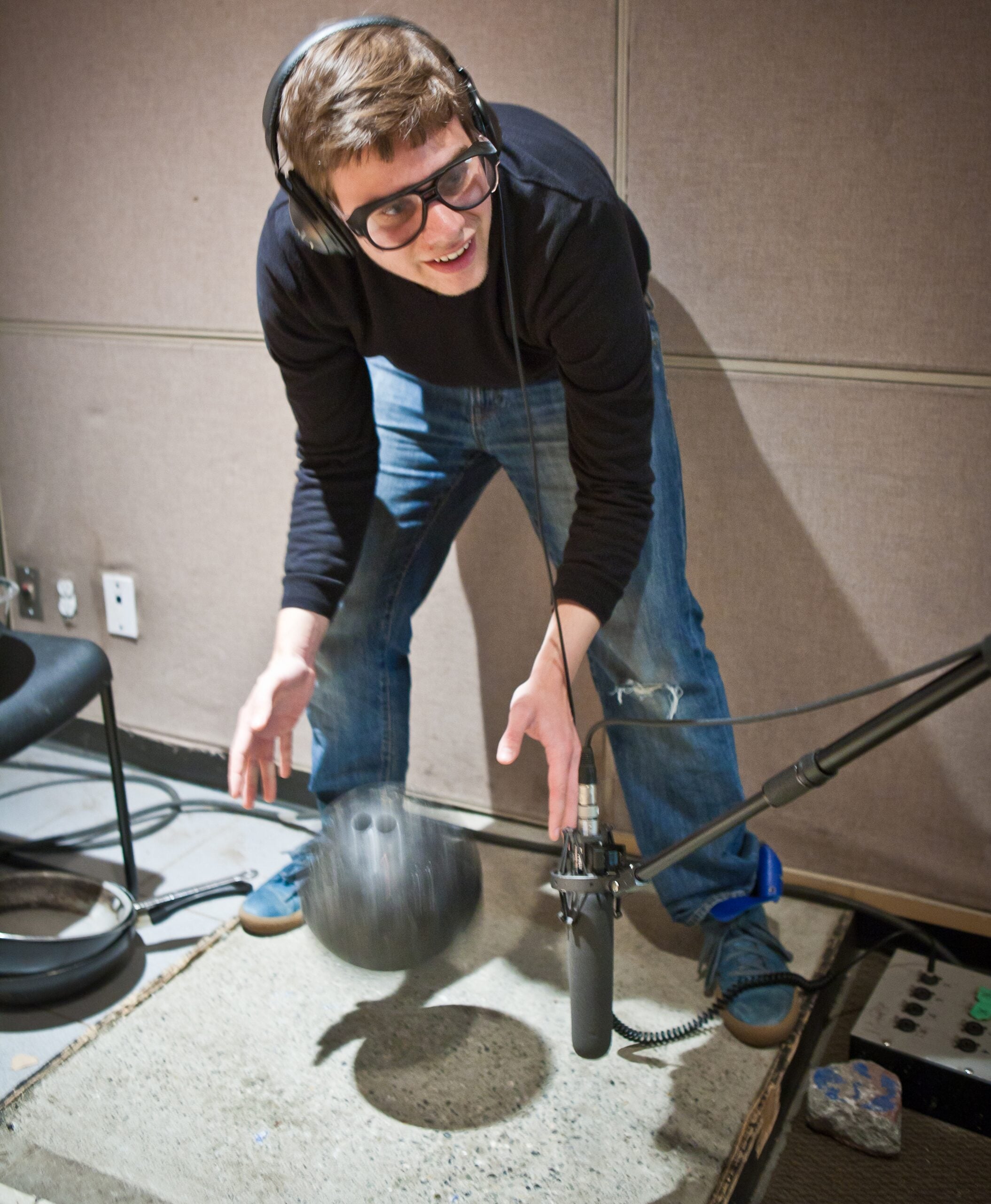 A person wearing headphones and glasses drops a bowling ball onto a concrete slab in a soundproof room, positioned near a microphone to capture the sound.