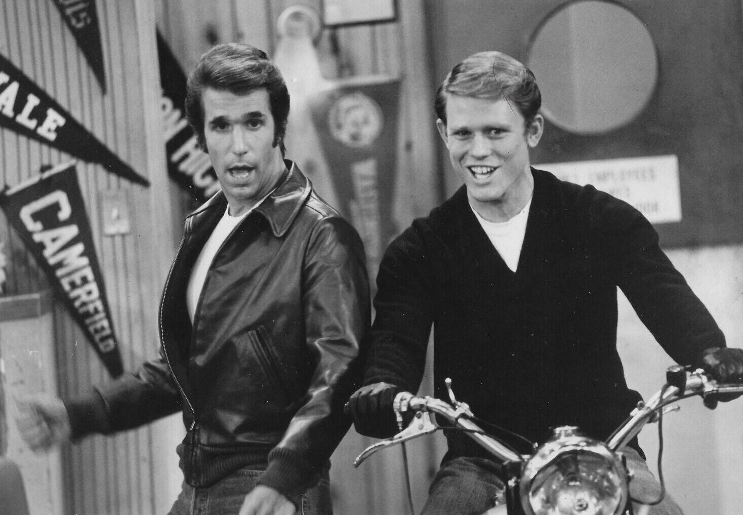 Two young men pose indoors; one wears a leather jacket, the other sits on a motorcycle and smiles. Pennants hang on the wall behind them. The scene has a vintage, black-and-white appearance.
