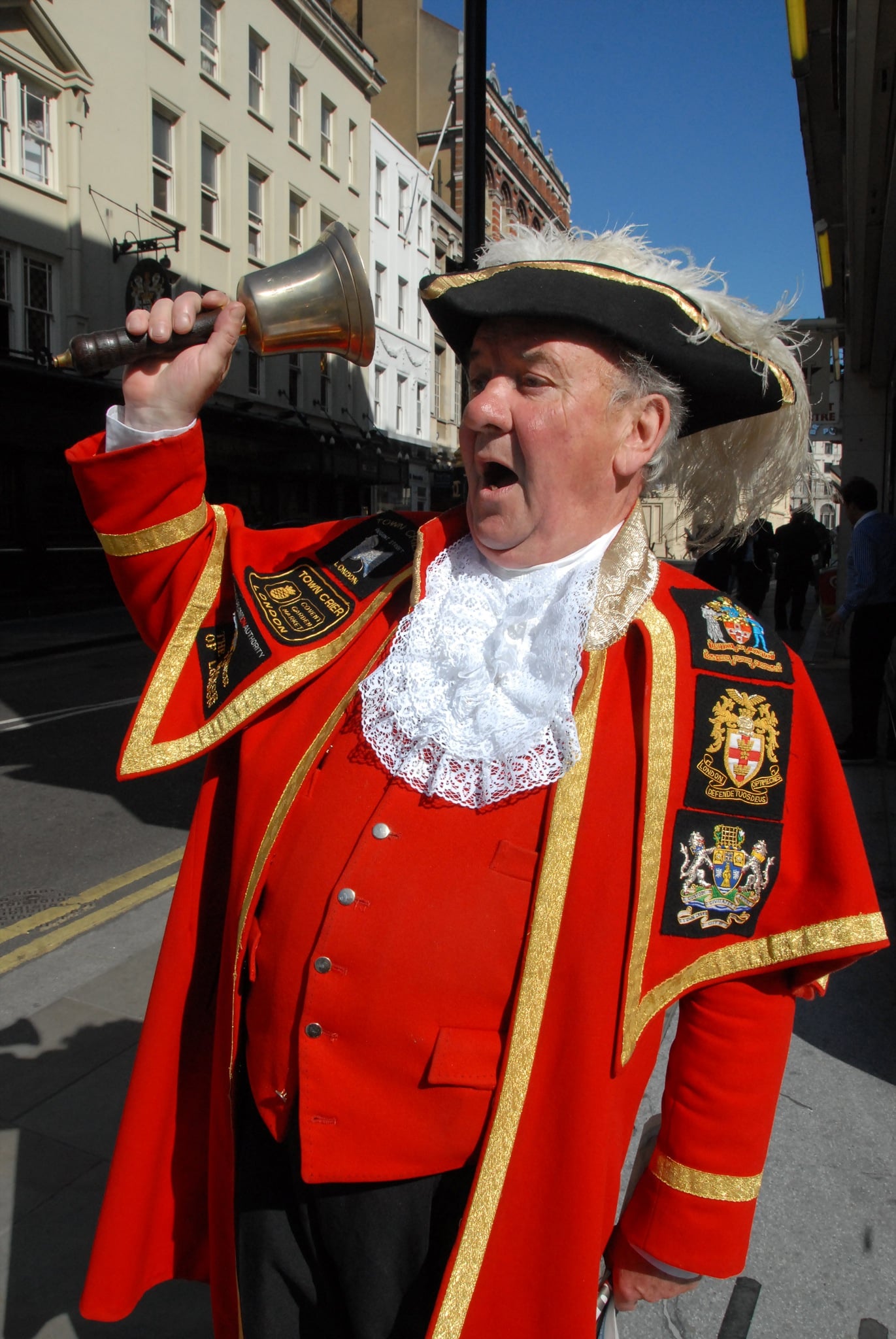A man dressed as a traditional town crier in a red coat, black hat with white trim, and lace ruff, holding a hand bell and calling out on a city street.