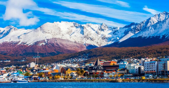 Colorful buildings line the waterfront with snow-capped mountains towering in the background under a bright blue sky.