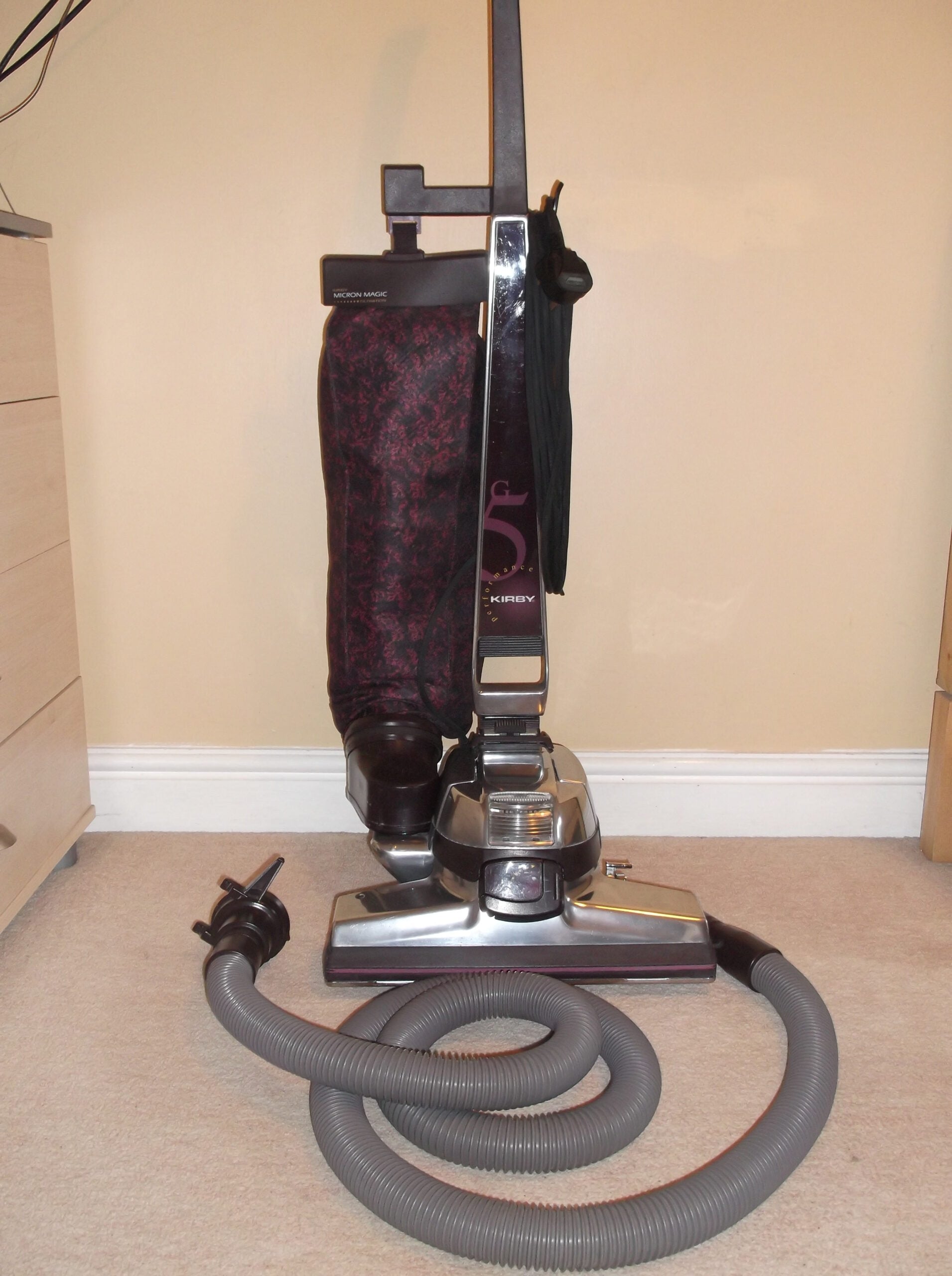 A vintage upright vacuum cleaner with an attached hose is positioned on a beige carpet in front of a cream-colored wall and next to a wooden dresser.