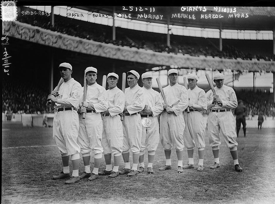 Nine baseball players in old-fashioned uniforms stand in a line on a field, some holding bats, with a large stadium and crowd in the background. Handwritten names appear at the top of the black and white photo.