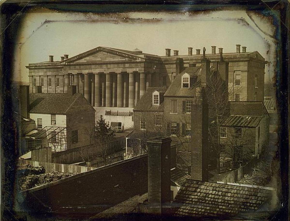 Sepia-toned photo of a large neoclassical building with tall columns behind smaller houses, chimneys, a greenhouse, trees, and a fenced yard in the foreground.