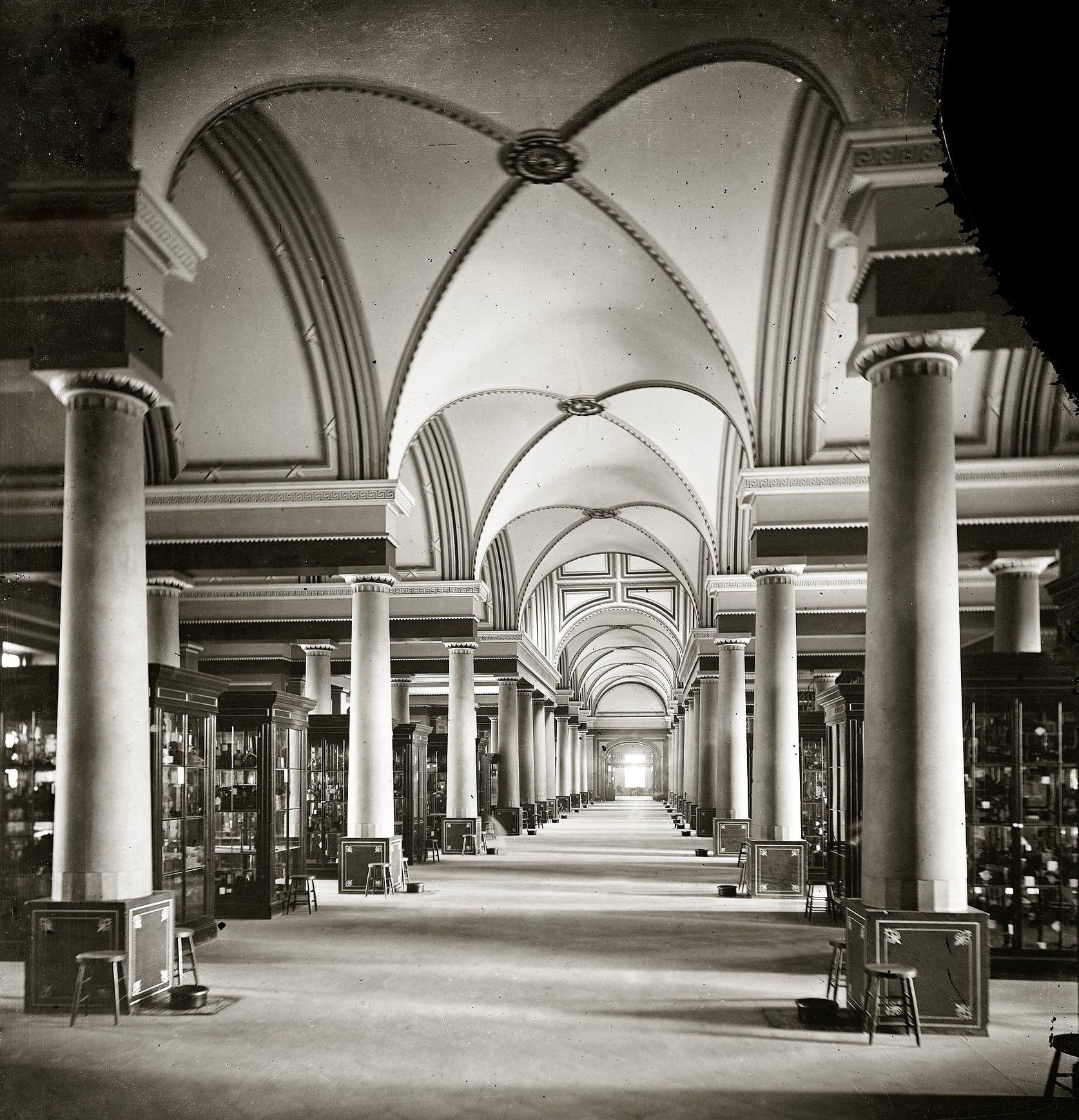 Grand hall with tall columns and arched vaulted ceilings, lined with glass display cases. The hall is spacious, well-lit, and symmetrical, creating a sense of depth and grandeur.