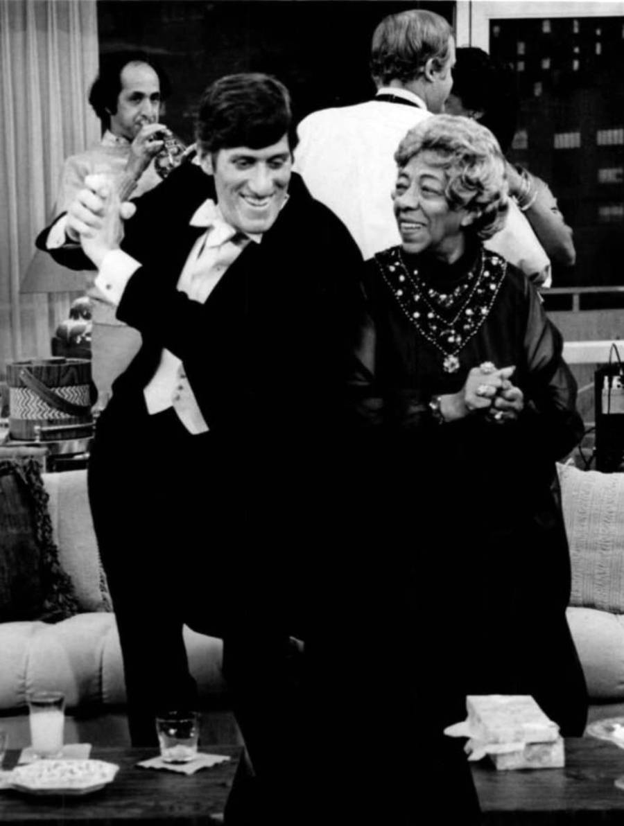 A smiling man in a tuxedo dances next to an older woman in a dress at a lively indoor gathering, with several people talking and mingling in the background.
