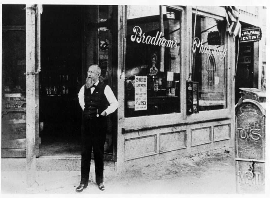 A man with a beard stands outside Bradham’s Pharmacy, an old-fashioned storefront with glass windows. A “U.S. Mail” mailbox is visible on the sidewalk in front of the store.