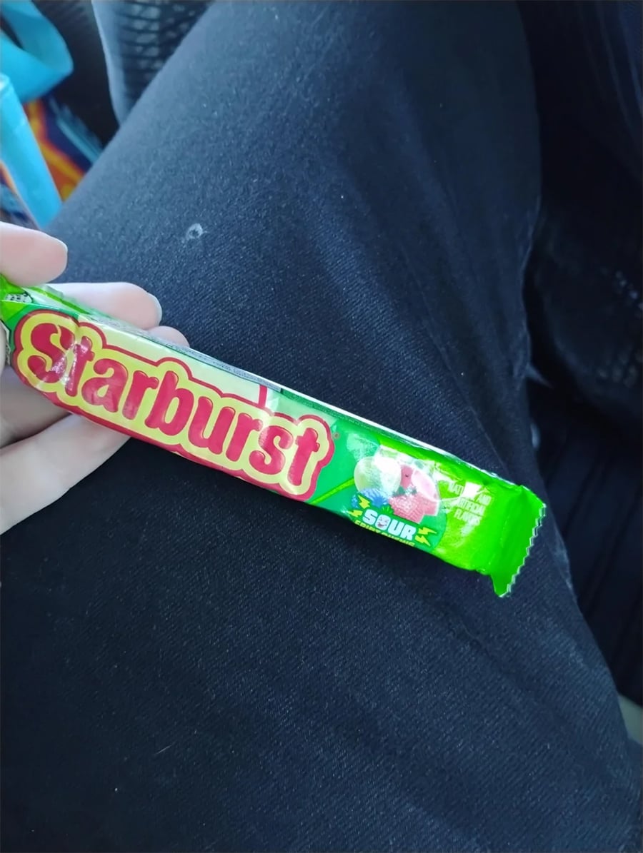 A hand holding a green package of Starburst sour candy, resting on a person’s black pants. The package design features images of the sour candies.