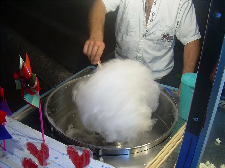 A person in a white shirt spins a large cloud of white cotton candy on a stick over a metal machine, with colorful pinwheels and red candy shapes visible nearby.