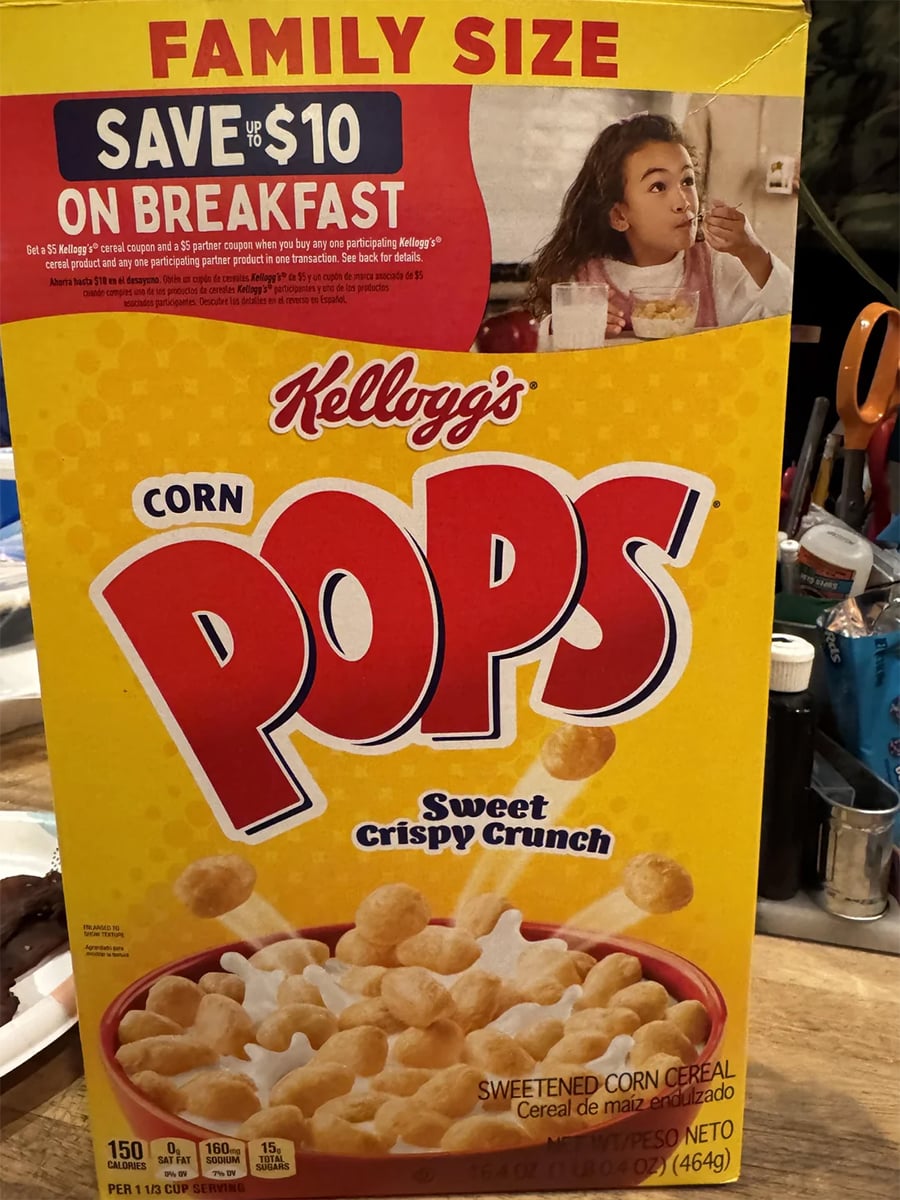 A family-size box of Kellogg’s Corn Pops cereal, featuring a bowl of the cereal and a child eating breakfast on the upper right corner. The box advertises a $10 breakfast savings offer.