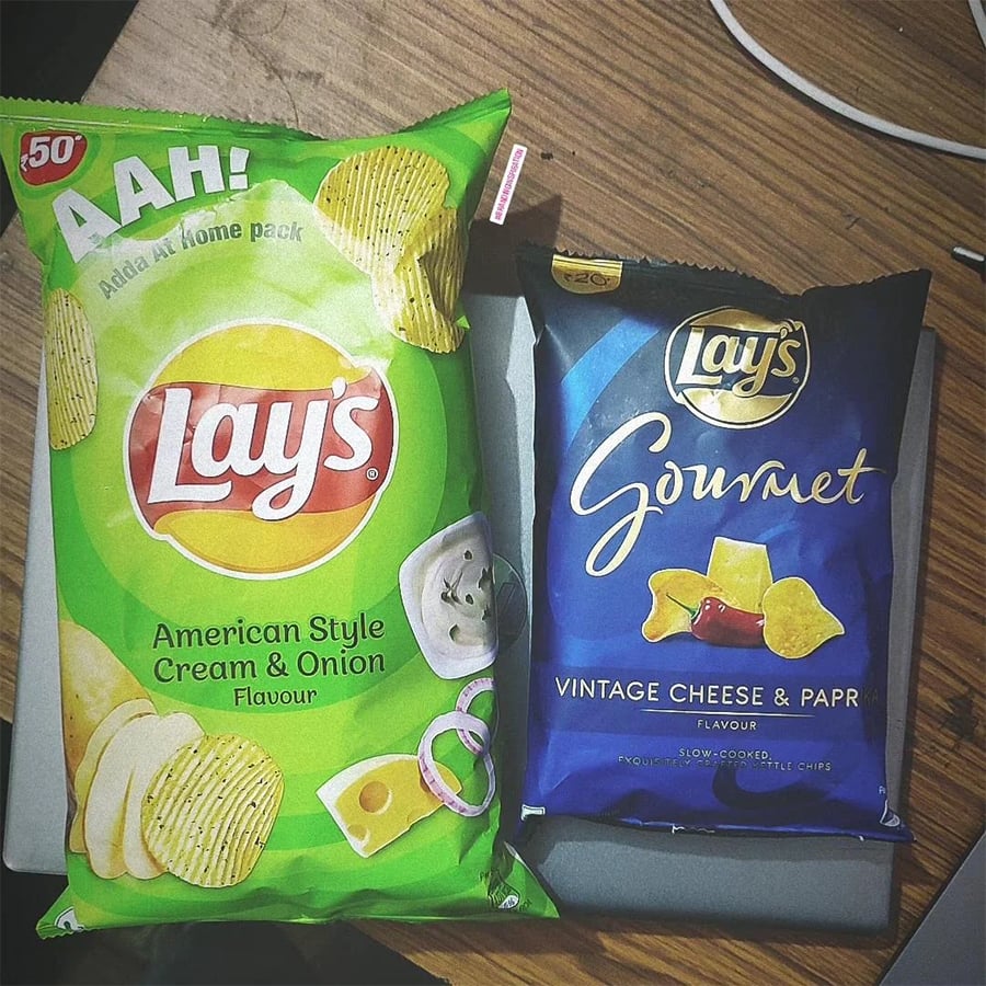 Two Lay’s potato chip bags on a wooden table: one green bag labeled “American Style Cream & Onion” and one blue bag labeled “Gourmet Vintage Cheese & Paprika.” Both bags are unopened and side by side.