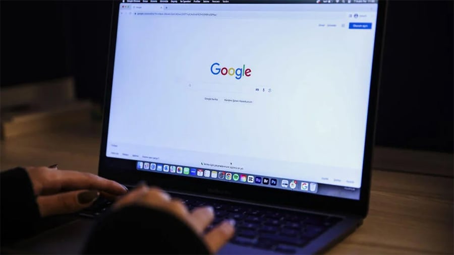 A person types on a laptop with the Google homepage open on the screen. The room appears dimly lit, and the focus is on the display and hands.