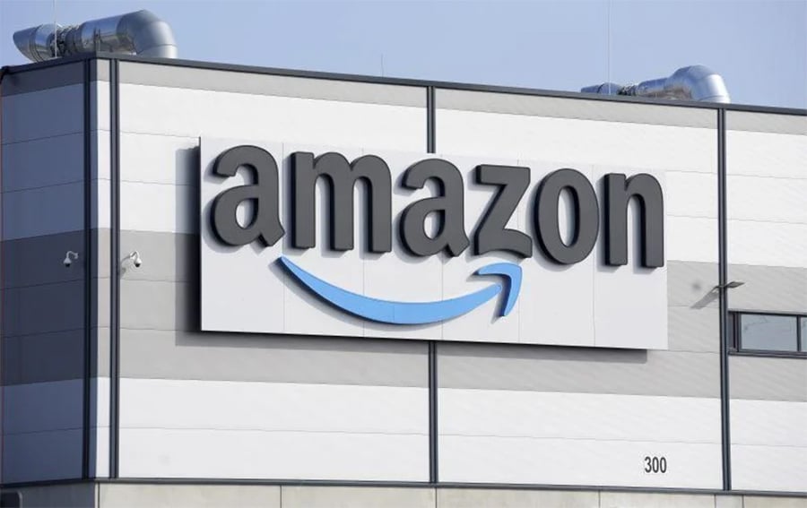 A large Amazon logo with an arrow underneath is displayed on the exterior wall of a modern warehouse building, with vents and a clear sky above.
