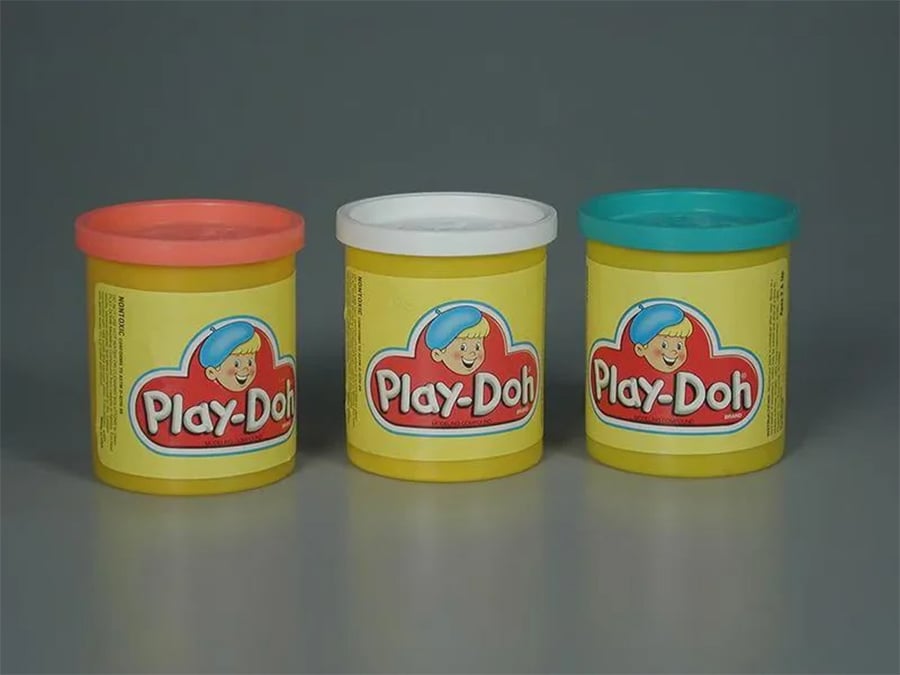 Three yellow Play-Doh containers are shown side by side with red, white, and blue-green lids. The label features a cartoon face and the Play-Doh logo on each container, against a plain gray background.