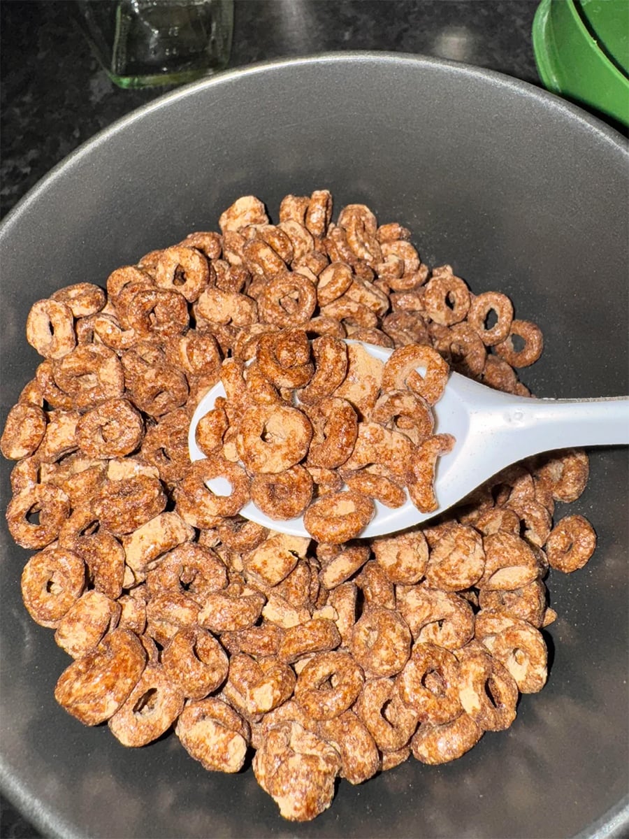 A gray bowl filled with chocolate-flavored cereal rings, with a white plastic spoon resting inside, ready to serve. The background is a dark kitchen countertop.