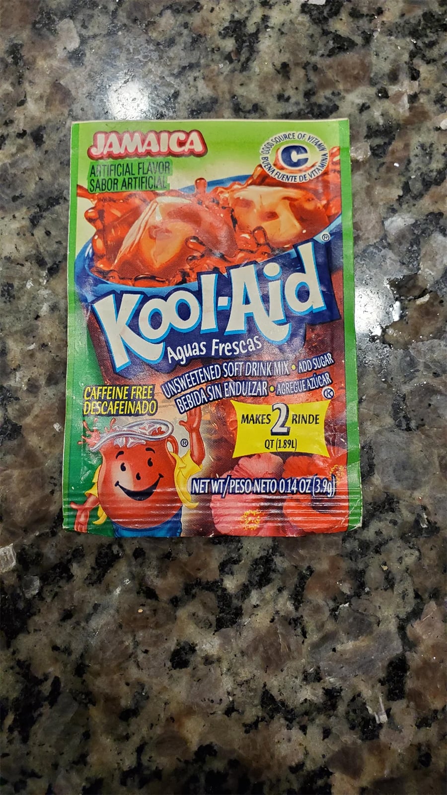 A Kool-Aid Aguas Frescas Jamaica drink mix packet lies on a granite countertop. The colorful packaging features a pitcher of red drink, the Kool-Aid Man, and instructions in English and Spanish.