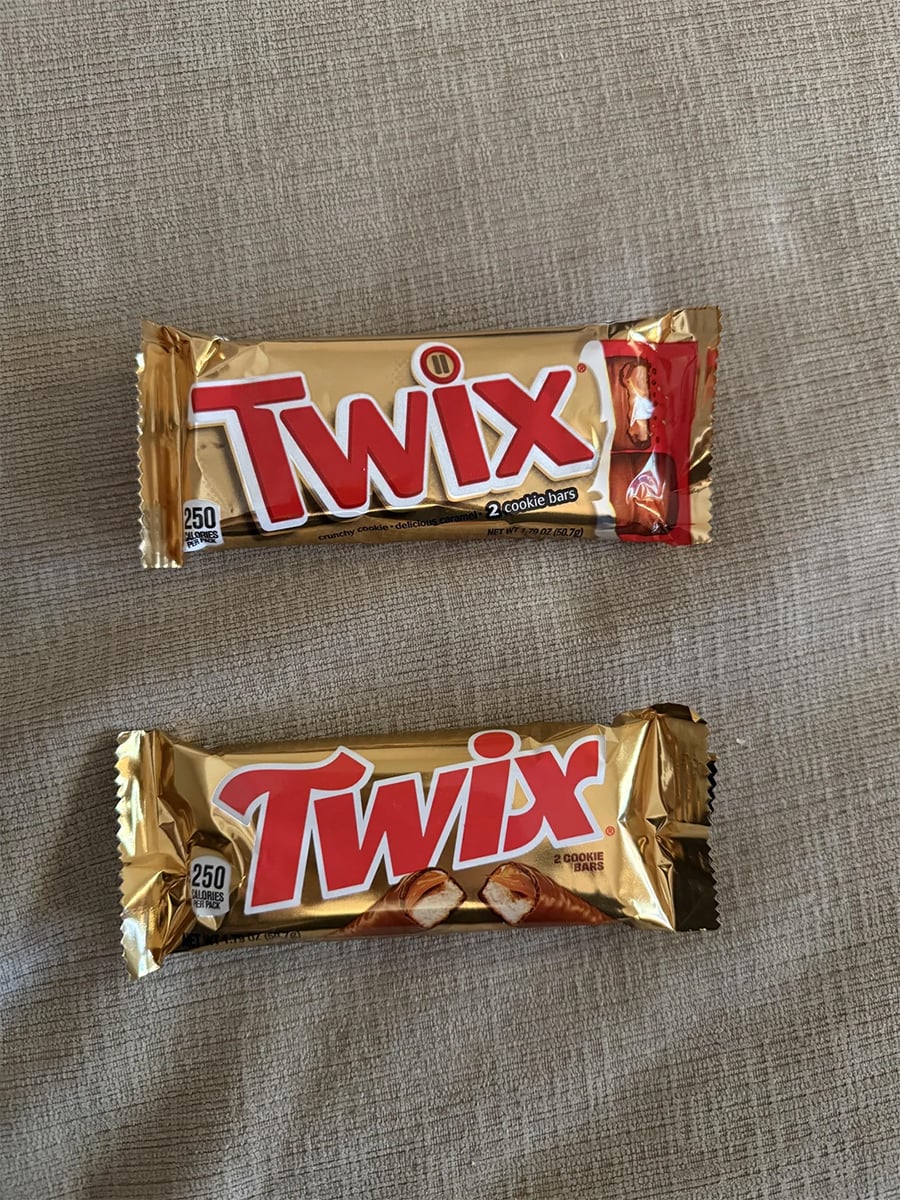 Two gold-wrapped Twix candy bars are on a beige fabric surface. The top package is larger and says “2 cookie bars,” while the bottom one is smaller and says “cookie bars.”