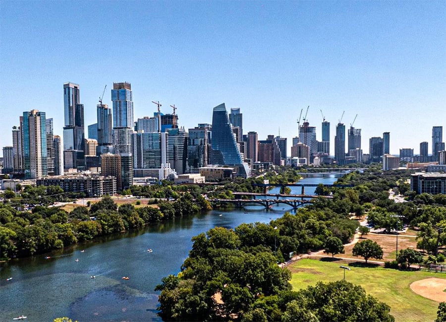 A city skyline with tall buildings, construction cranes, and a river running through a park with trees and a baseball field under a clear blue sky.