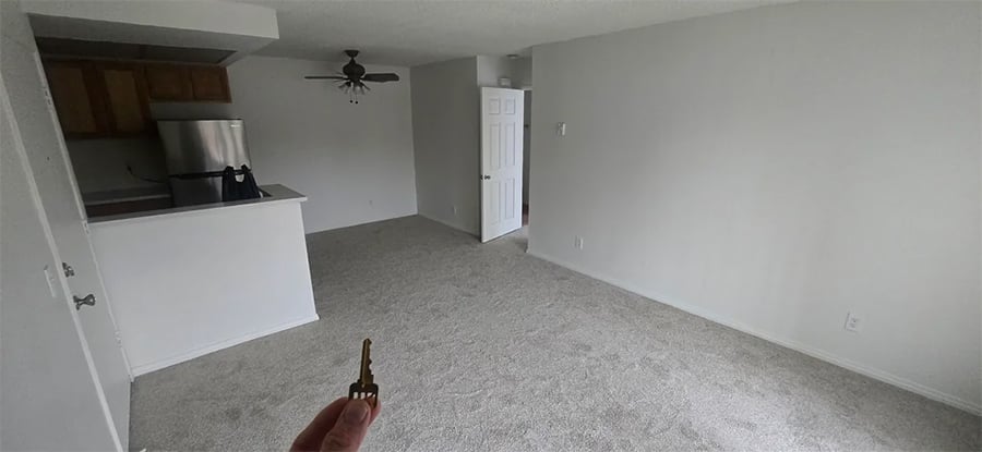 A person holds a key in an empty apartment with carpeted floors, white walls, a small kitchen area, a ceiling fan, and an open door leading to another room.