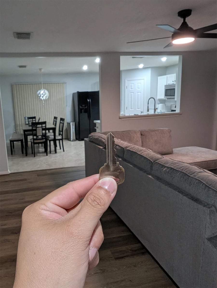 A hand holding a key in a living room with a gray sectional sofa. In the background, there is a dining table, kitchen, refrigerator, and ceiling fan with lights.