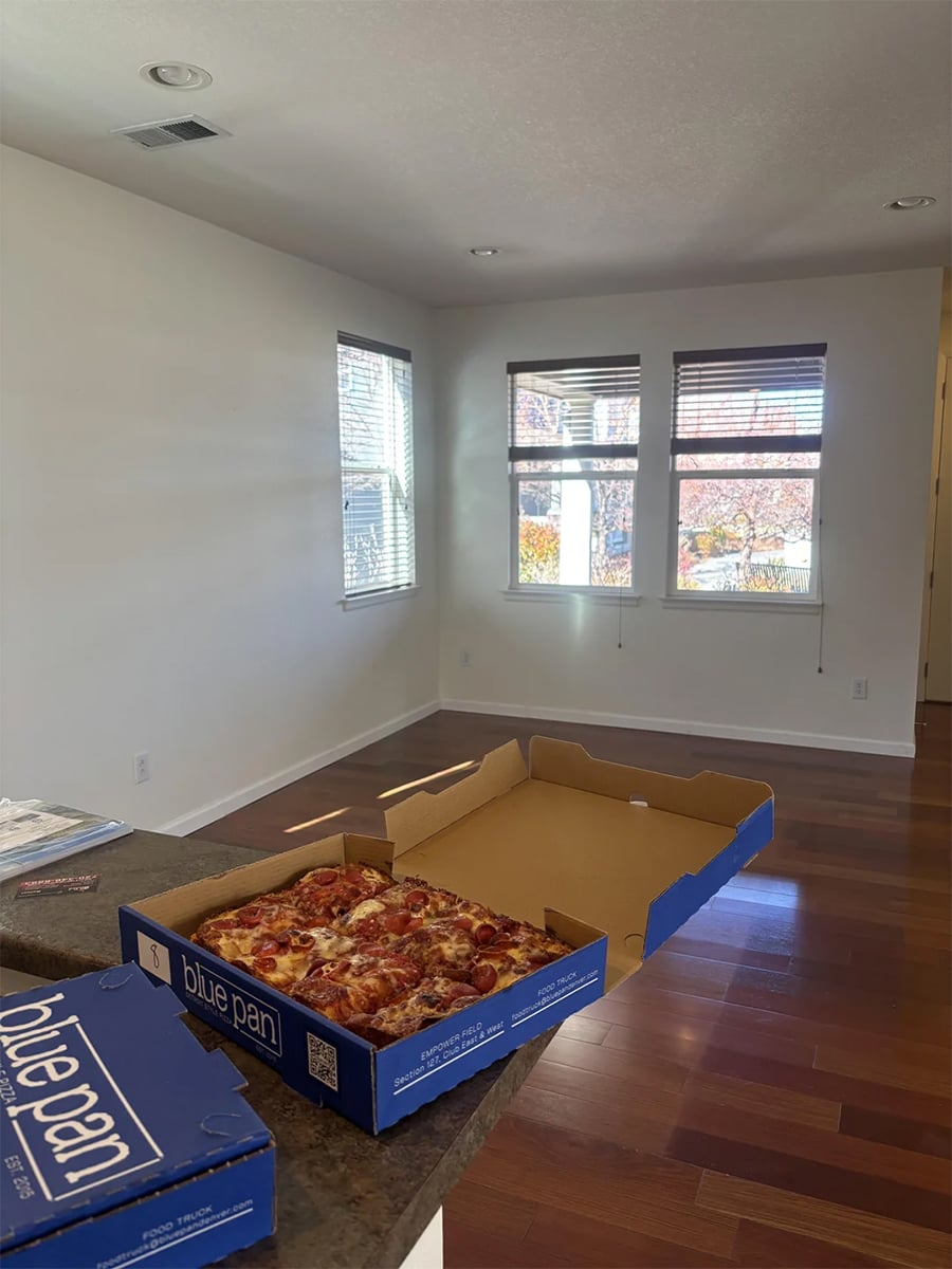 An open pizza box with pizza sits on a kitchen counter in a mostly empty room with wooden floors and large windows letting in sunlight.