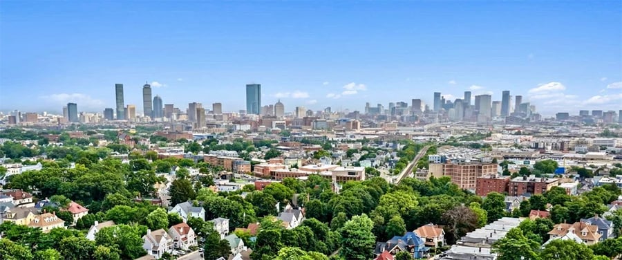 A panoramic view of a city skyline under a clear blue sky, with tall skyscrapers in the distance and residential houses and green trees in the foreground.