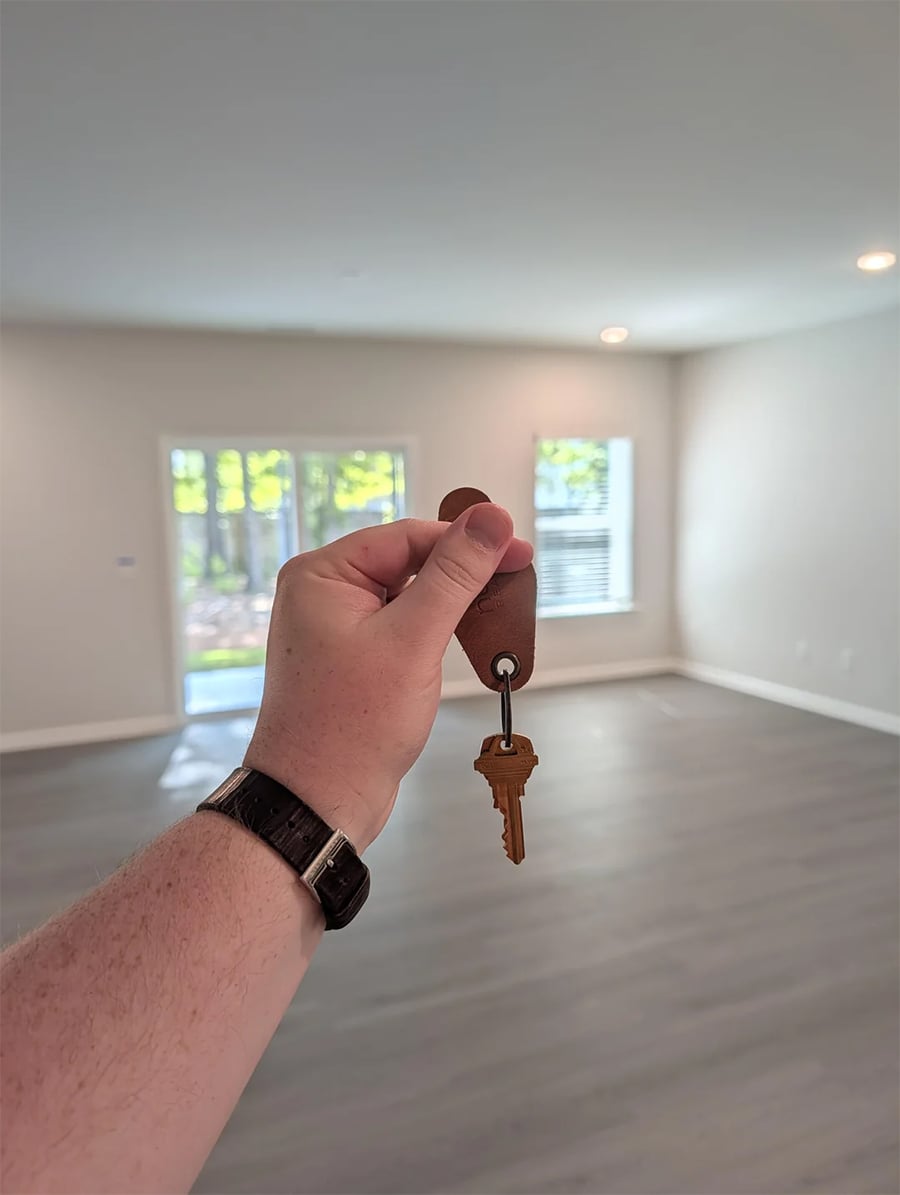 A hand holding up a house key in an empty, well-lit room with large windows and light-colored floors.