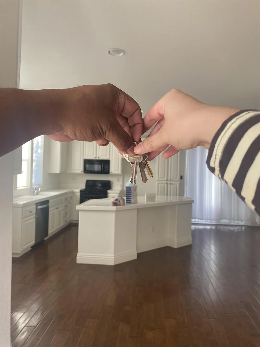 Two hands, one darker and one lighter, hold a set of keys together in front of a modern, empty kitchen with white cabinets, wooden floors, and natural light.
