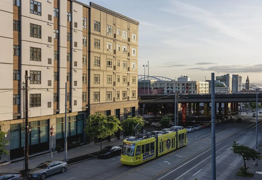 A yellow streetcar travels down a city street beside a modern apartment building, with cars parked along the curb and an overpass and city skyline visible in the background.