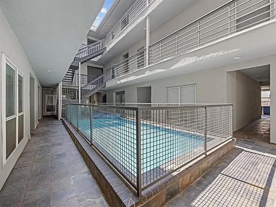 A narrow indoor swimming pool is surrounded by metal railings and tiled flooring, with walkways and stairs on two levels under a partial skylight.