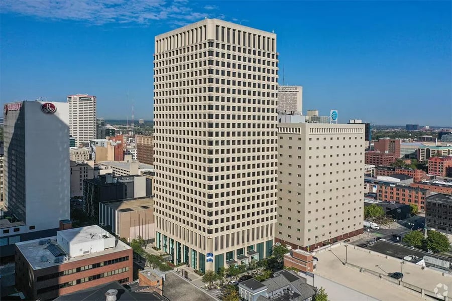 A tall, beige office building stands in a cityscape surrounded by other buildings under a clear blue sky, with streets and some greenery visible at the base.