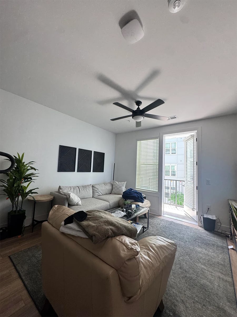 A bright living room with a beige couch, coffee table, indoor plants, wall art, and an open door leading to a balcony. A ceiling fan and light fixtures are on the white ceiling.