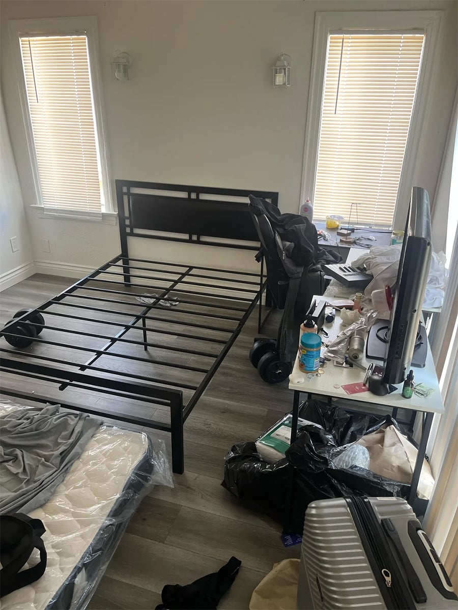 A messy bedroom with an empty black bedframe, an unwrapped mattress on the floor, blinds on windows, cluttered desk with a TV, suitcase, trash bags, and various items scattered around.