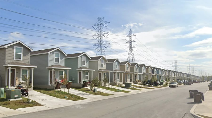A row of modern two-story houses lines a suburban street, with power lines and tall transmission towers visible in the background under a partly cloudy sky.