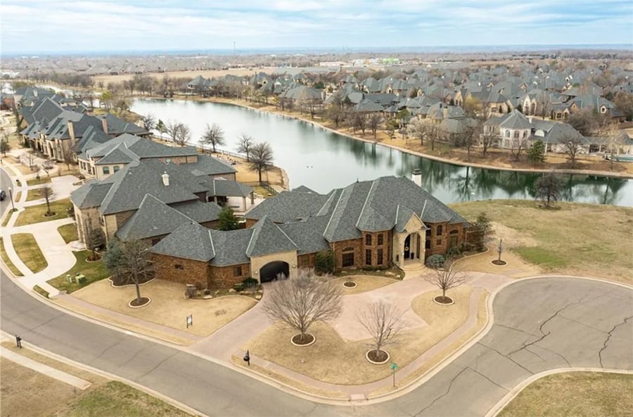 Aerial view of a large brick house on a corner lot by a lake, surrounded by similar homes with gray roofs in a suburban neighborhood.