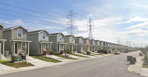 A row of modern, identical two-story houses lines a suburban street, with cars parked in driveways and tall electrical towers and power lines stretching overhead.