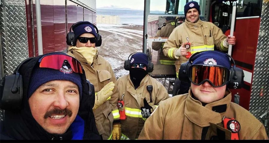 Five firefighters in tan gear and winter hats stand near a fire truck on a snowy day. Some wear goggles and headsets; one smiles in front for a selfie. Buildings and fields are visible in the background.