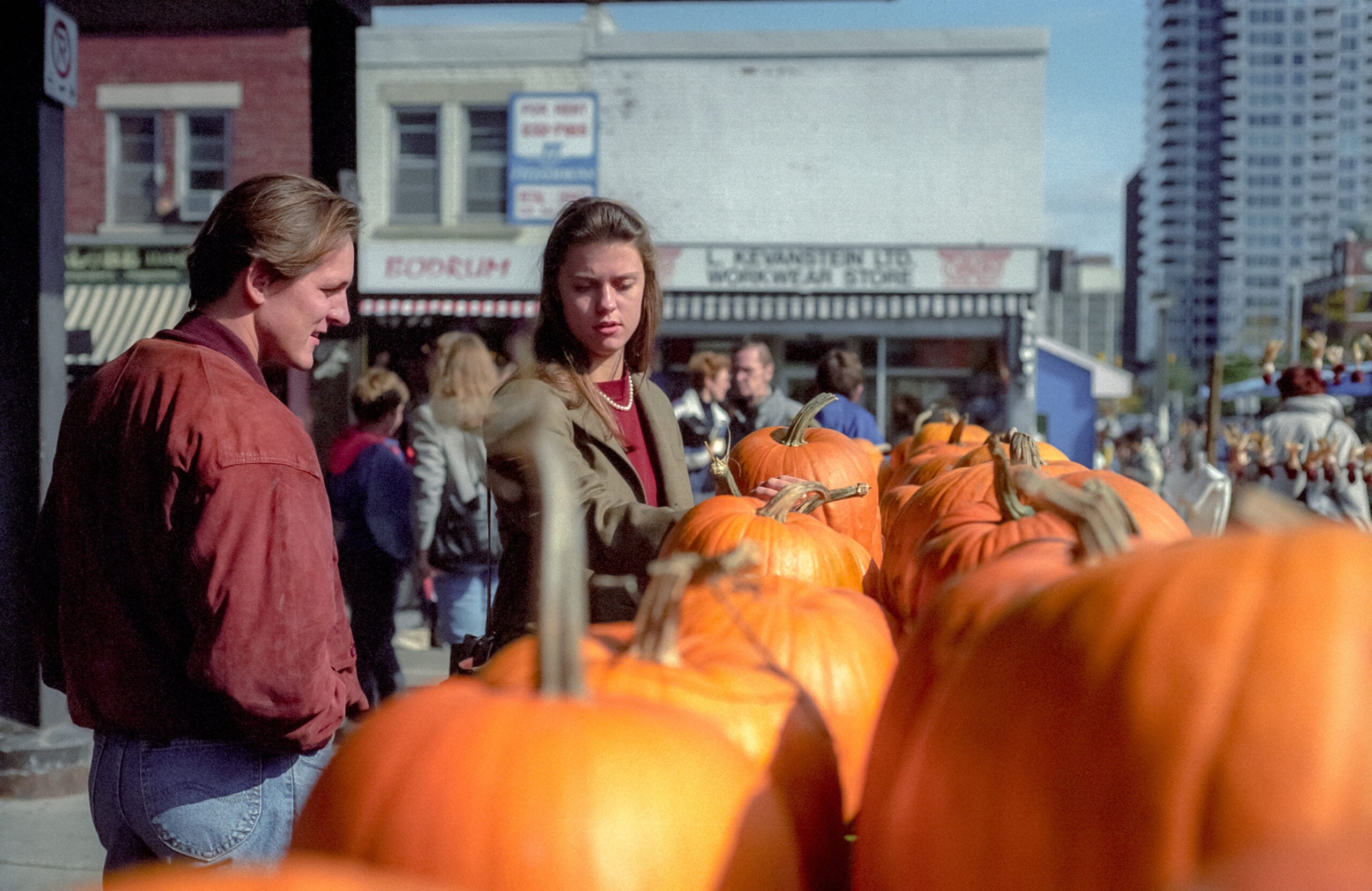 Two people stand by a display of large pumpkins at an outdoor market, with other shoppers and city buildings visible in the background.