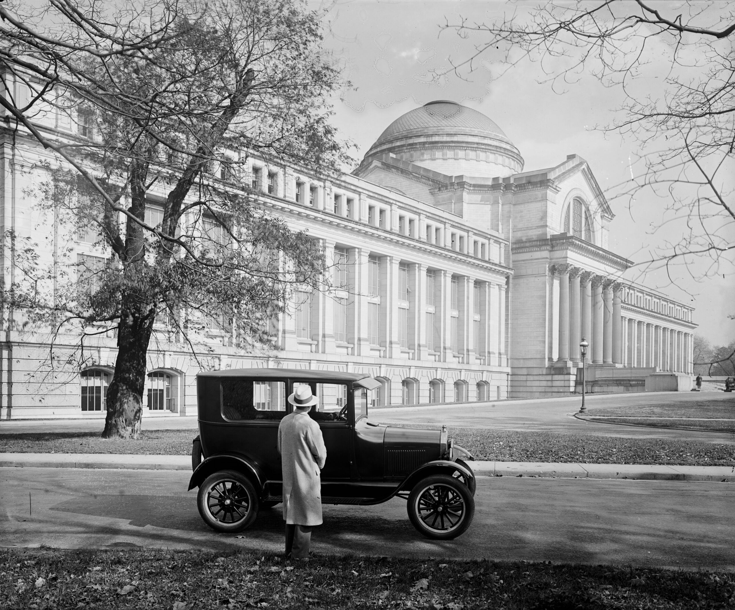 A person in a coat and hat stands beside a vintage car, facing a large neoclassical building with columns and a domed roof on a tree-lined street.