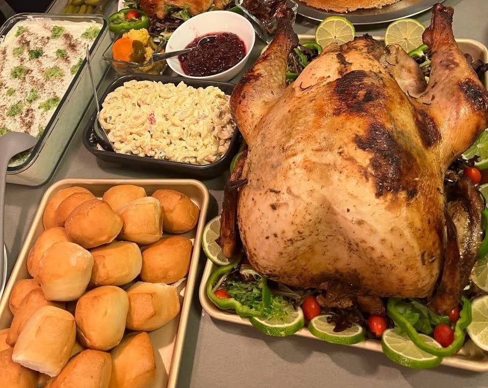 A roasted turkey on a platter with lime slices and greens, surrounded by dinner rolls, pasta salad, cranberry sauce, and a vegetable casserole on a table.