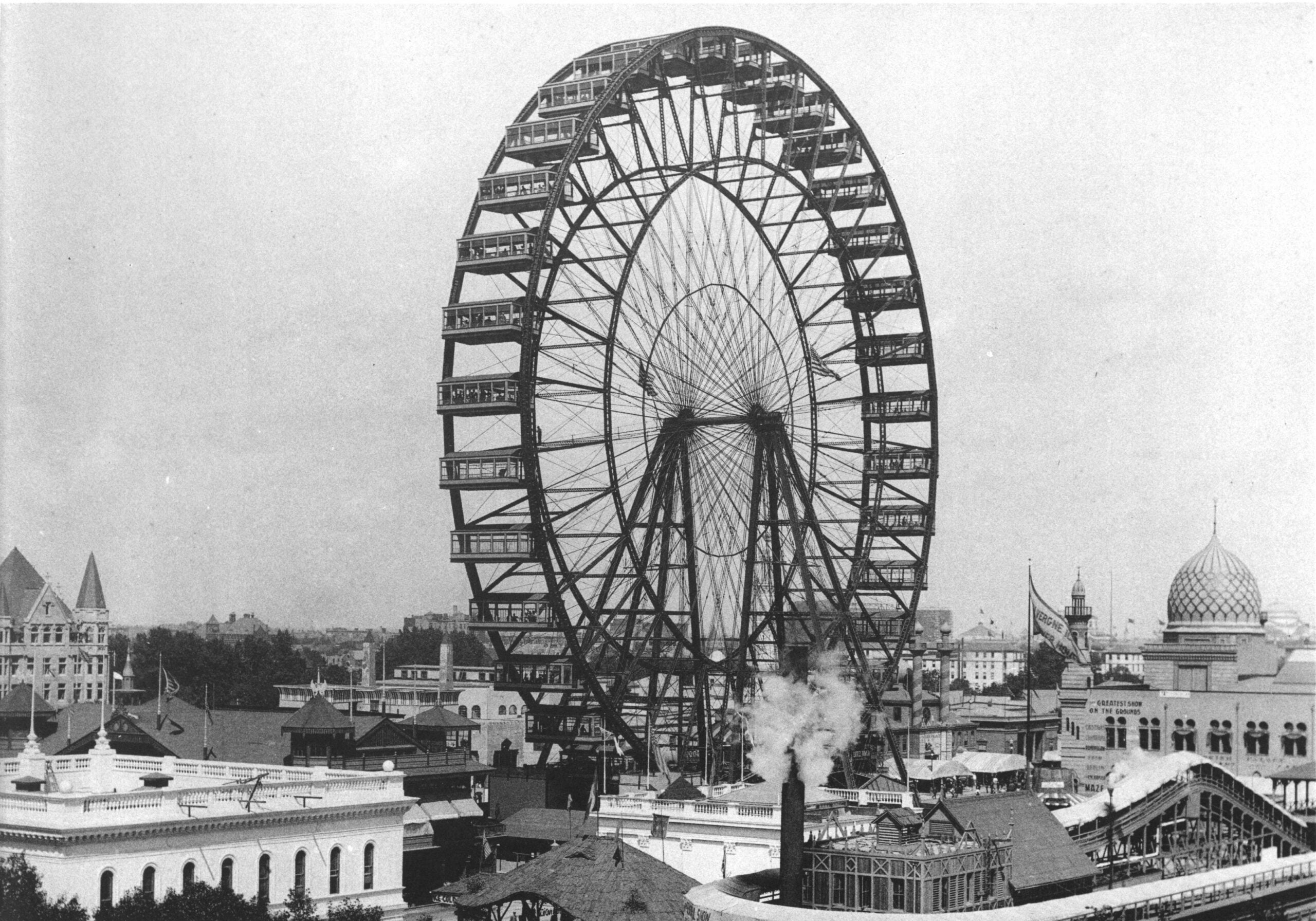 Black and white photo of a large Ferris wheel towering over buildings at a fairground. The area is bustling with structures, some with domes, and a smokestack releases steam in the foreground.